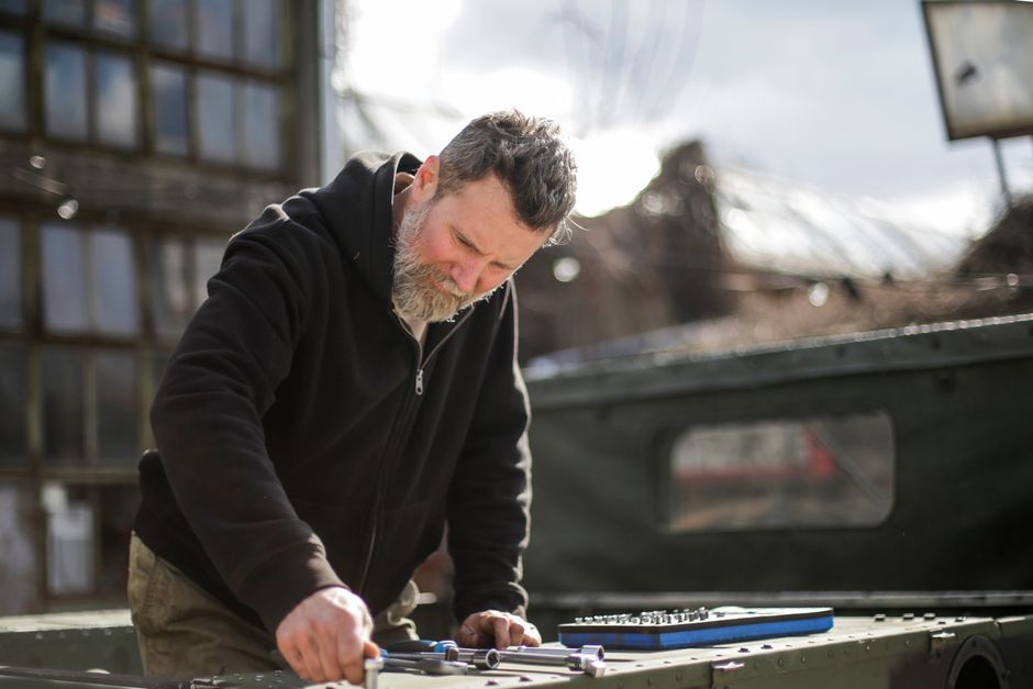Serious adult bearded mechanic in workwear standing on trunk of car and checking details with ratchet while working with toolbox on street near garage